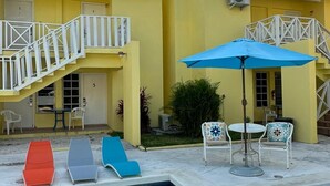 A pool area with lounge chairs and a table with chairs, surrounded by a yellow building with a staircase and an air conditioning unit. - Sun Fun Resort (Nassau)