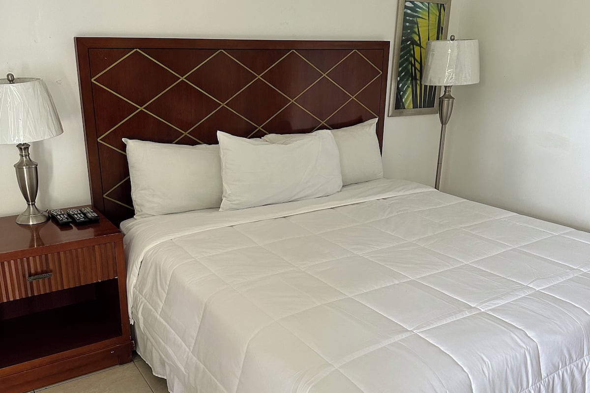 Una cama bien hecha con ropa blanca, un cabecero de madera con diseño de diamante, una mesita de noche con lámpara y controles remotos, y un cuadro en la pared.