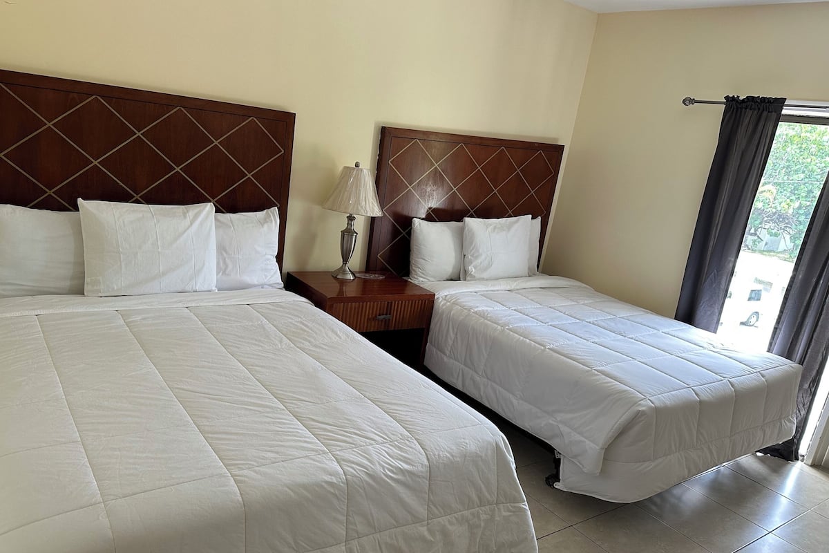 Habitación de hotel con dos camas, cada una con cabecera de madera, ropa de cama blanca y mesita de noche con lámpara.