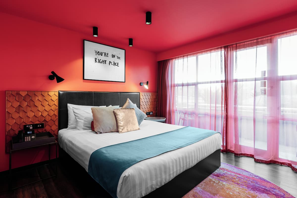Een moderne hotelkamer met een groot bed, een houten hoofdbord, een nachtkastje met een telefoon en een raam met dunne gordijnen.