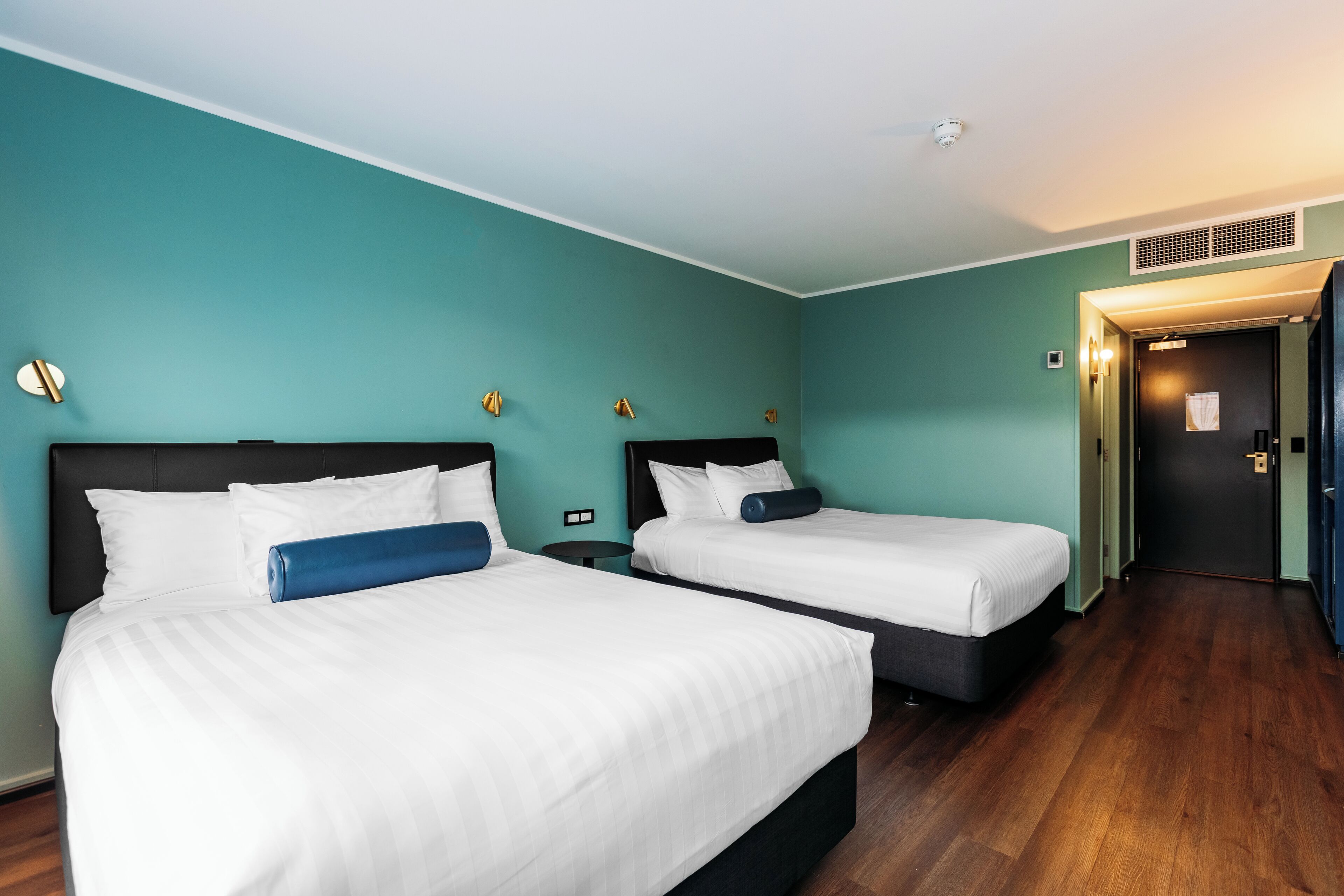 Quarto de hotel com duas camas, paredes verde-água e soalho de madeira.