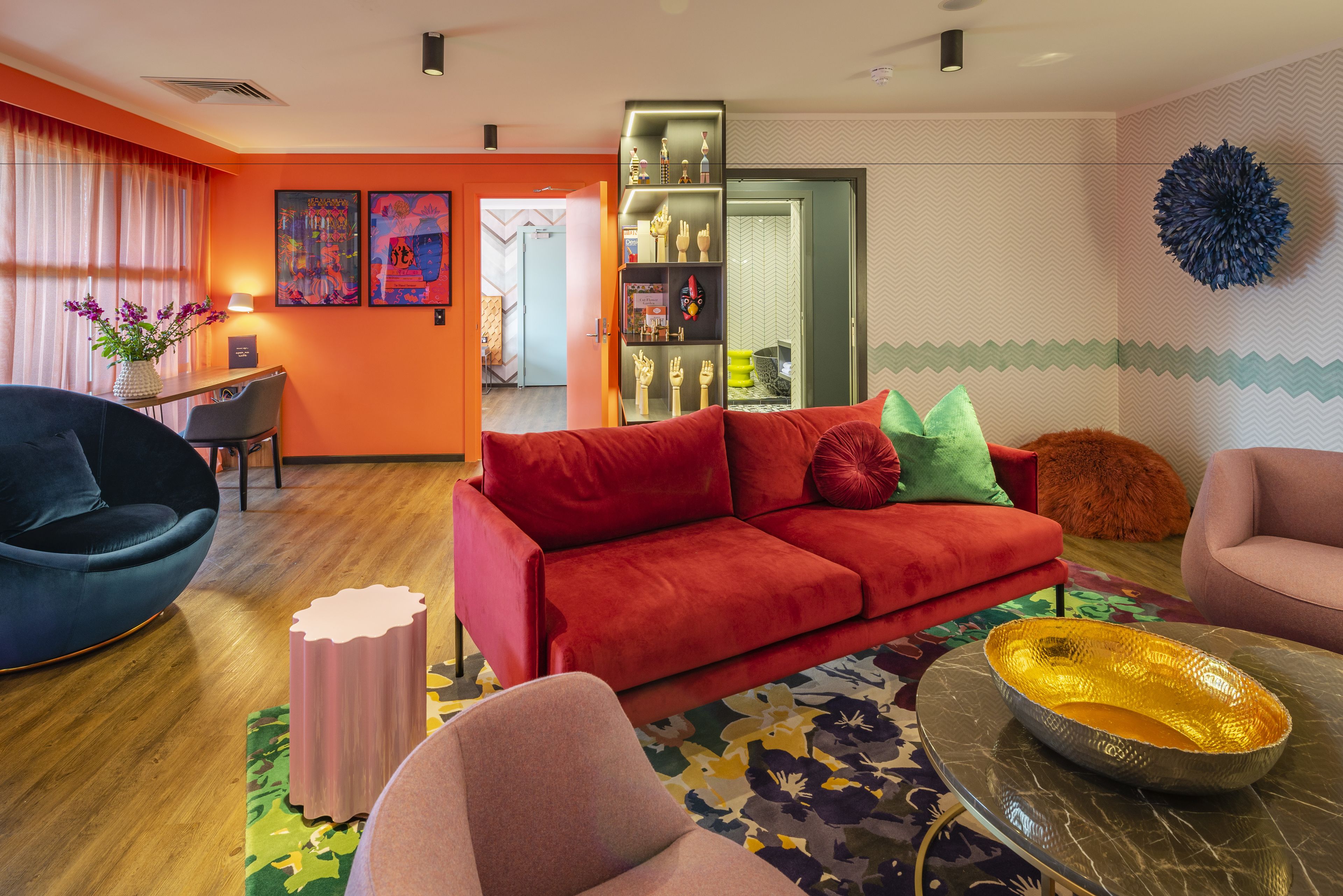 Una sala de estar moderna con un sofá rojo, un sillón negro y un sillón rosa. Hay una mesita con un florero y una lámpara. Se ve una estantería con objetos decorativos al fondo.