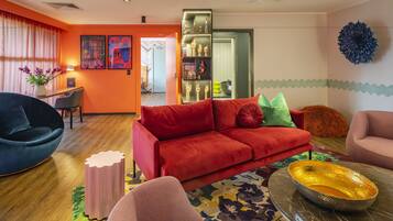 A modern living room with a red sofa, a black armchair, and a pink armchair. There is a small table with a vase of flowers and a lamp. A bookshelf with decorative items is visible in the background.