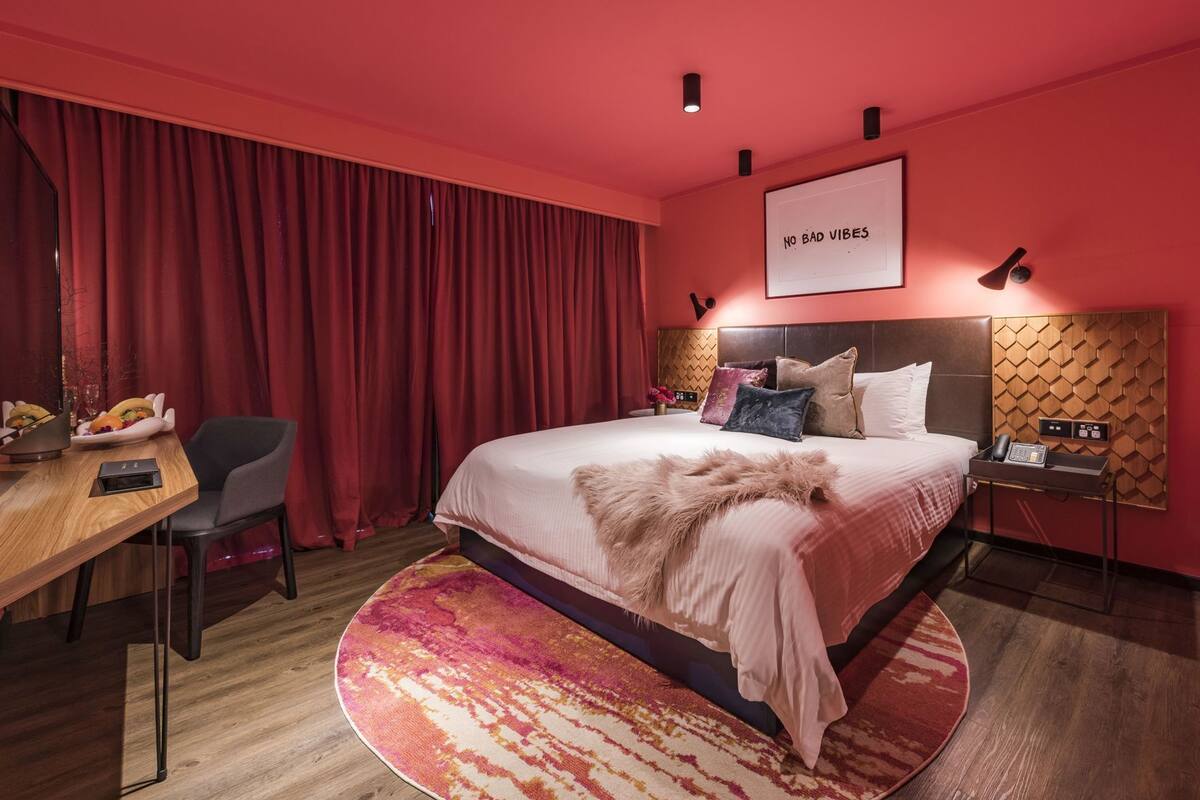 Une chambre d’hôtel avec un grand lit, un bureau, une chaise, un rideau rouge, un téléphone fixé au mur et une pancarte sur laquelle on peut lire : « PAS D’ÉNERGIES NÉGATIVES ».