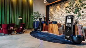 Un espace d’accueil d’hôtel au design moderne, comprenant un comptoir d’accueil en bois, un kiosque d’information numérique et des fauteuils rouges recouverts de tissu.