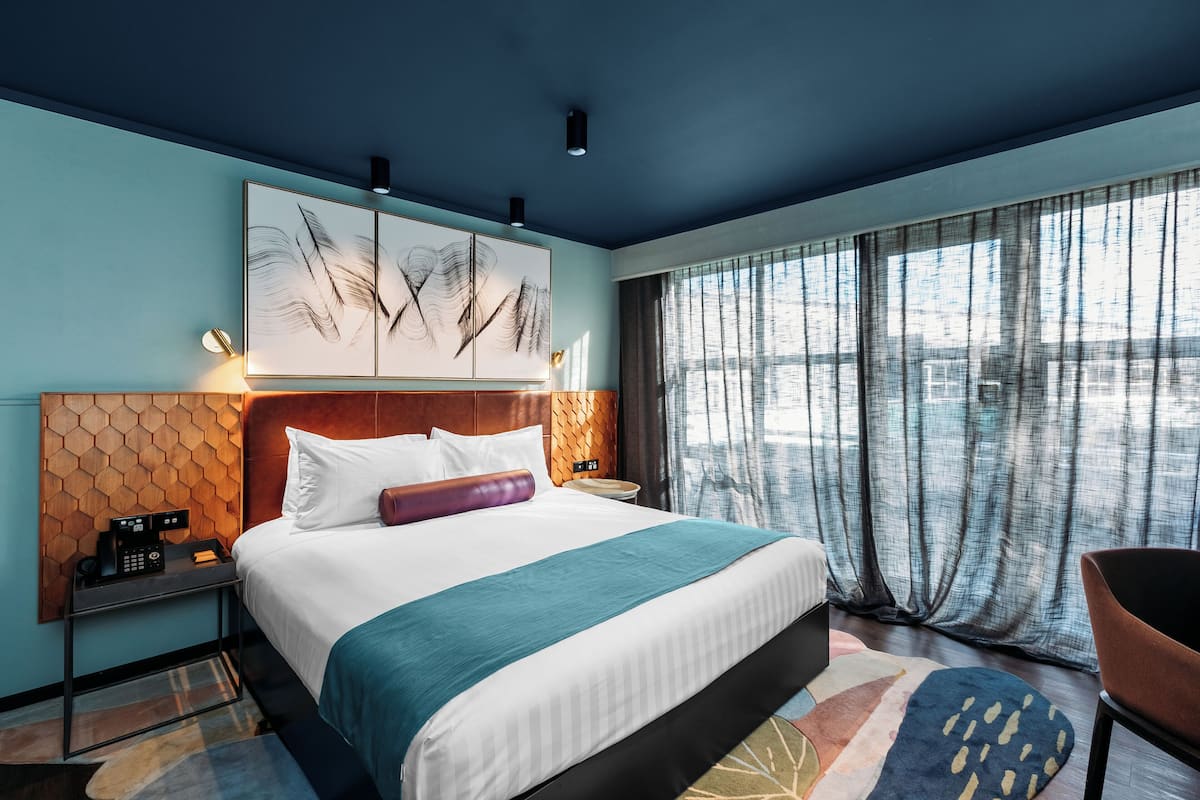 Een moderne hotelkamer met een groot bed, een houten hoofdbord, een telefoon aan de muur en uitzicht op de stad door dunne gordijnen.