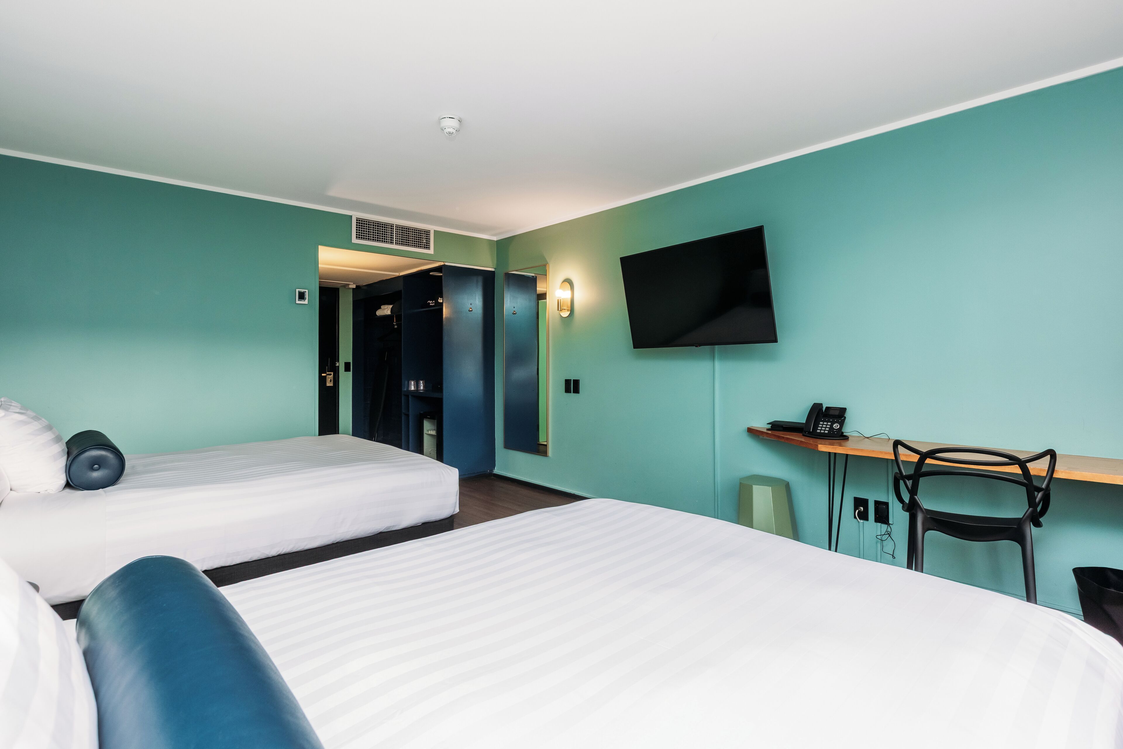 Une chambre d’hôtel avec deux lits, un bureau, une chaise, une télévision et une petite poubelle.
