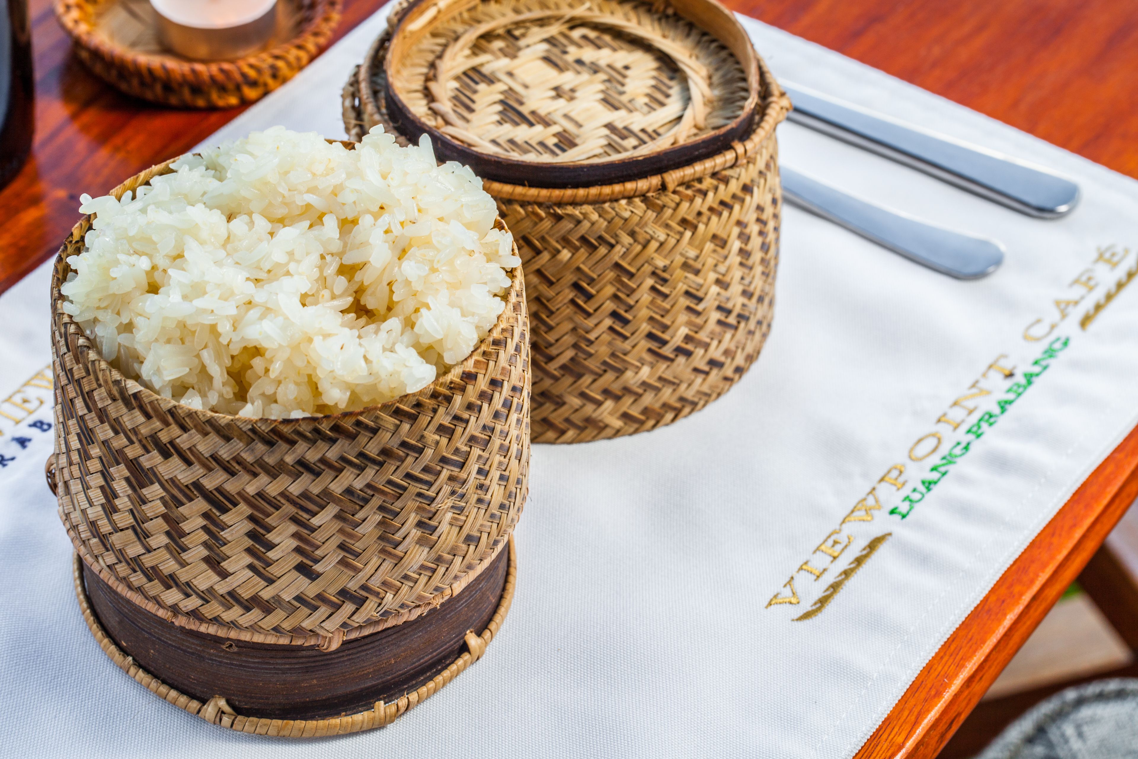 Two woven bamboo containers filled with white rice, placed on a table with a placemat that has the text "VIEWPOINT LUANG PRABONG" and a pair of silver utensils.