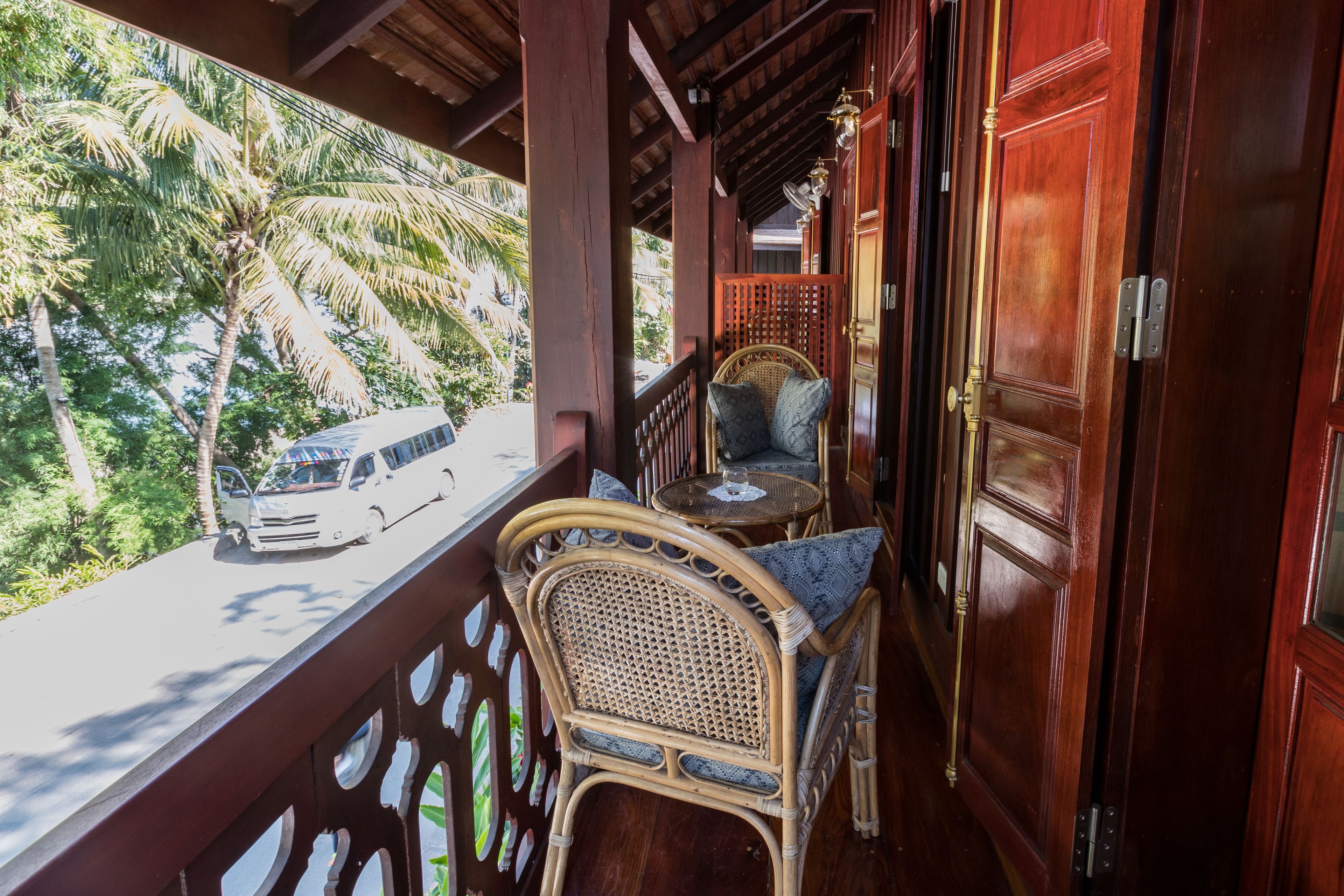 A balcony with a wicker chair and a small table, overlooking a street with a parked car and palm trees.
