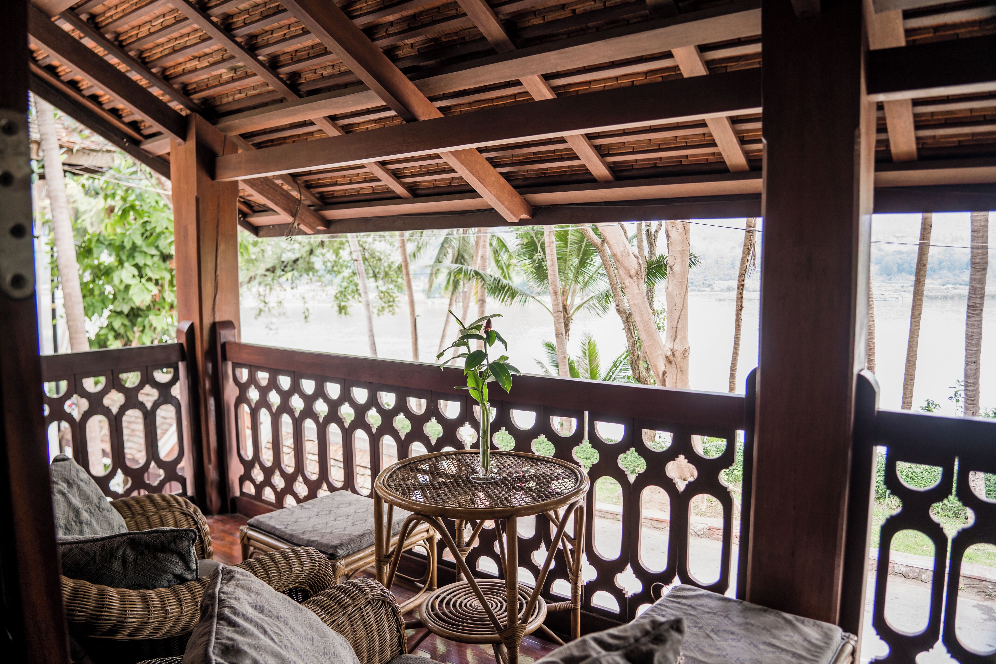 A covered outdoor seating area with wicker furniture and a central round table, overlooking a beach with palm trees.