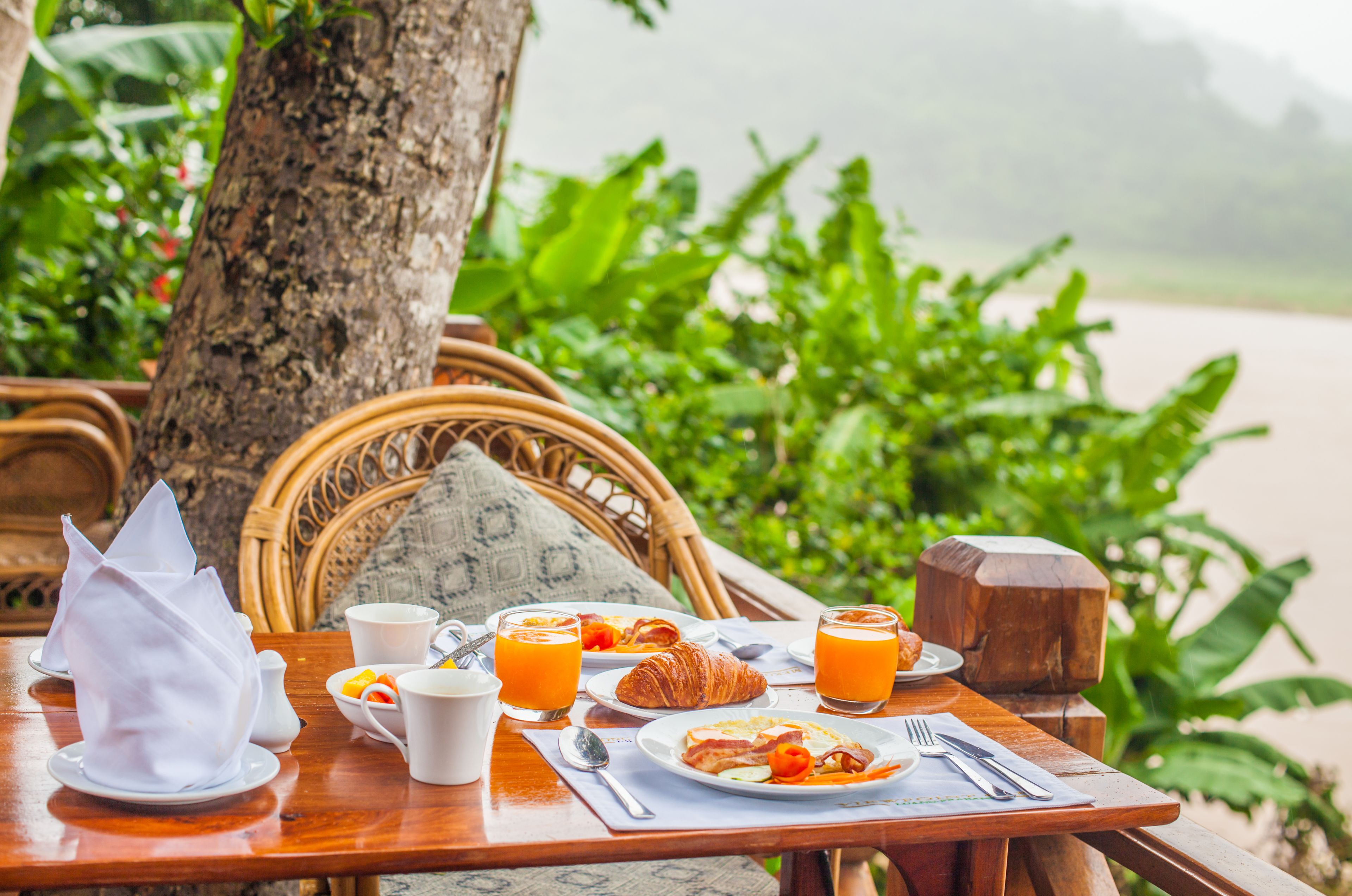 A breakfast table with croissants, orange juice, and coffee cups, set outdoors with a wicker chair and a tree in the background.