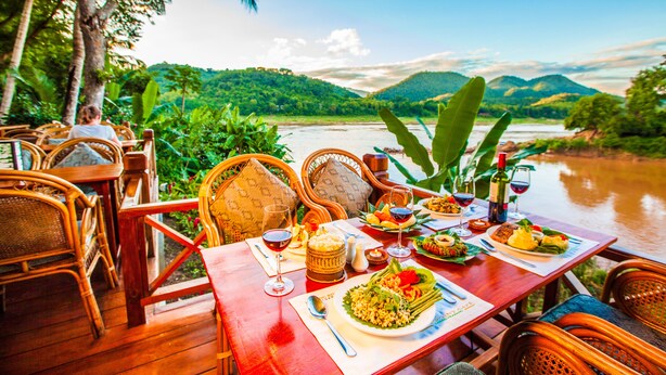 Une table à manger dressée avec des assiettes remplies de nourriture, des verres à vin et une bouteille de vin, donnant sur une rivière et une végétation luxuriante.
