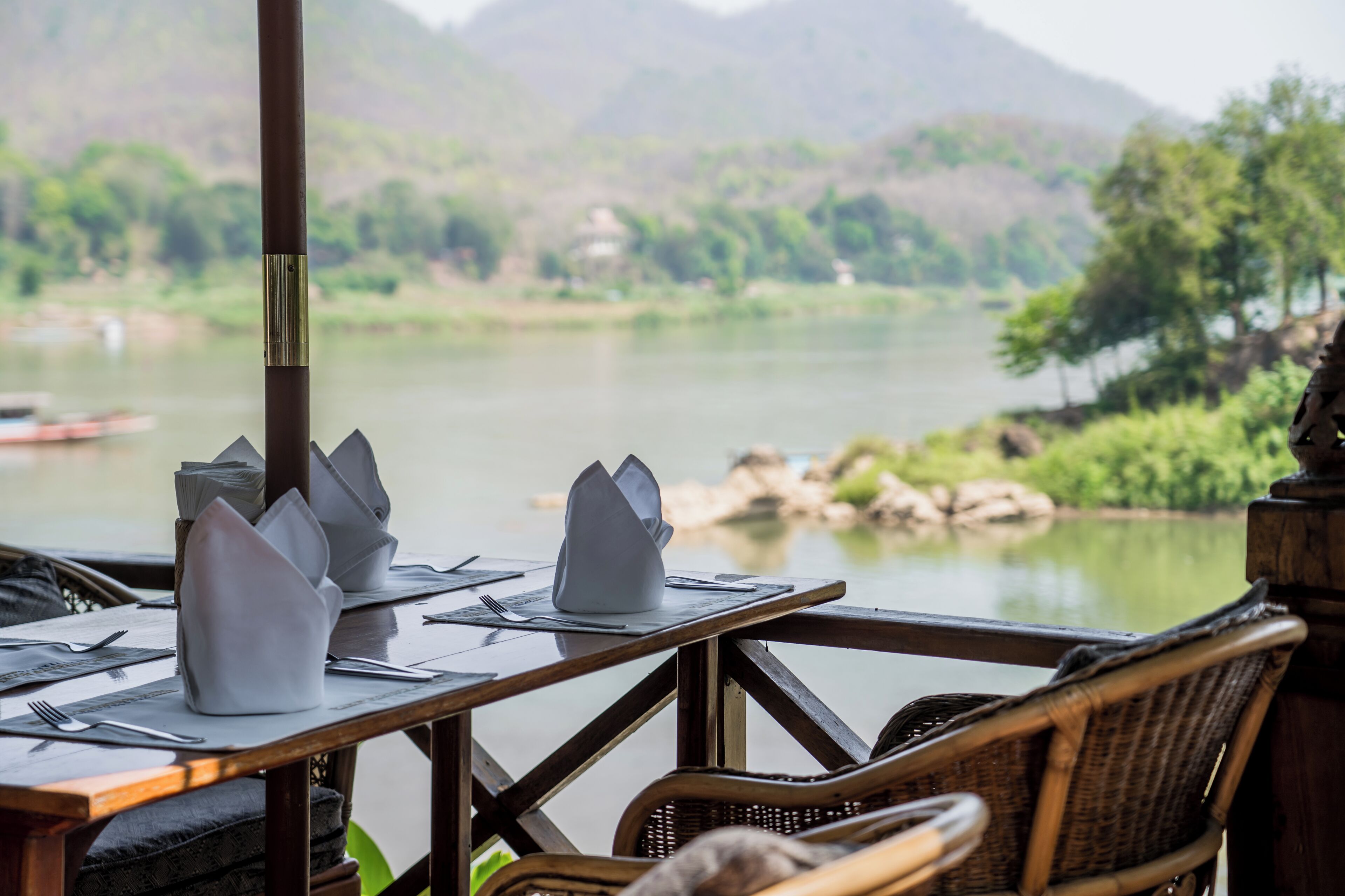 Mesa de comedor dispuesta con servilletas y cubiertos blancos, con vista a un río tranquilo y exuberante follaje.