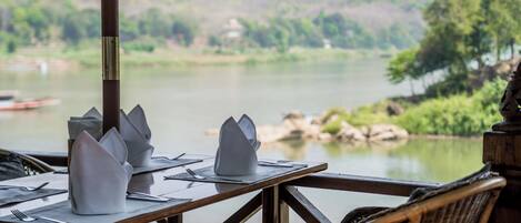 Mesa de comedor dispuesta con servilletas y cubiertos blancos, con vista a un río tranquilo y exuberante follaje.
