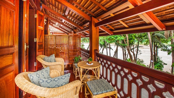 A wooden balcony with wicker furniture, a view of palm trees, and a wooden lattice partition. - Mekong Riverview Hotel (Luang Prabang)