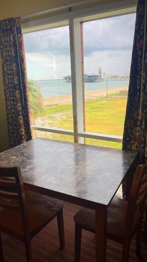 A room with a large window overlooking a beach and a ship. There is a table with chairs in the foreground.