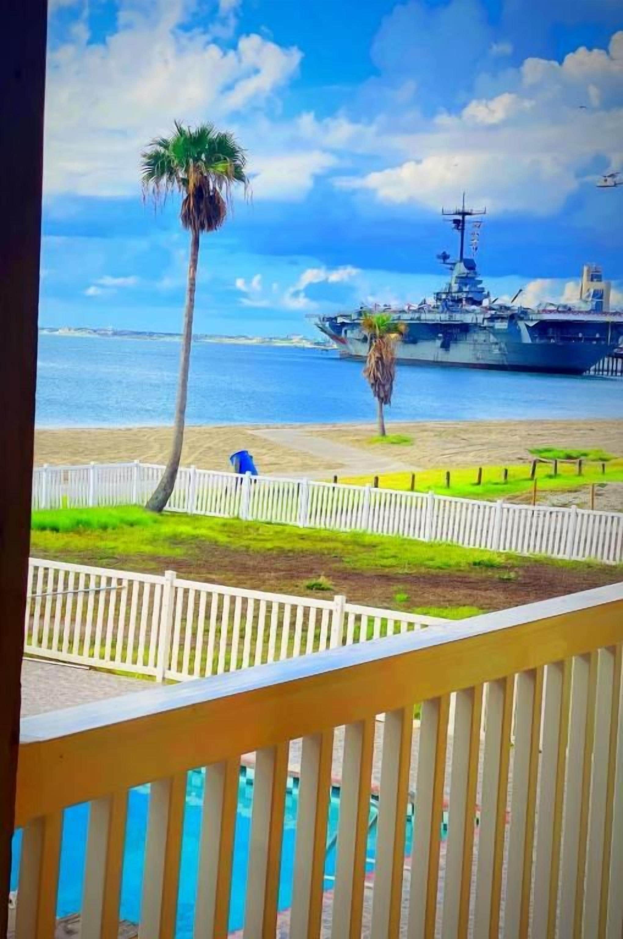 Vista de una playa con un barco militar al fondo, una palmera y una cerca blanca.