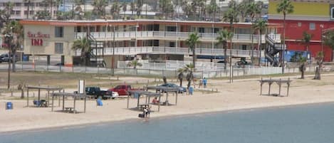Un hotel frente a la playa con varios balcones, piscina y zona de playa con algunas personas y coches aparcados.