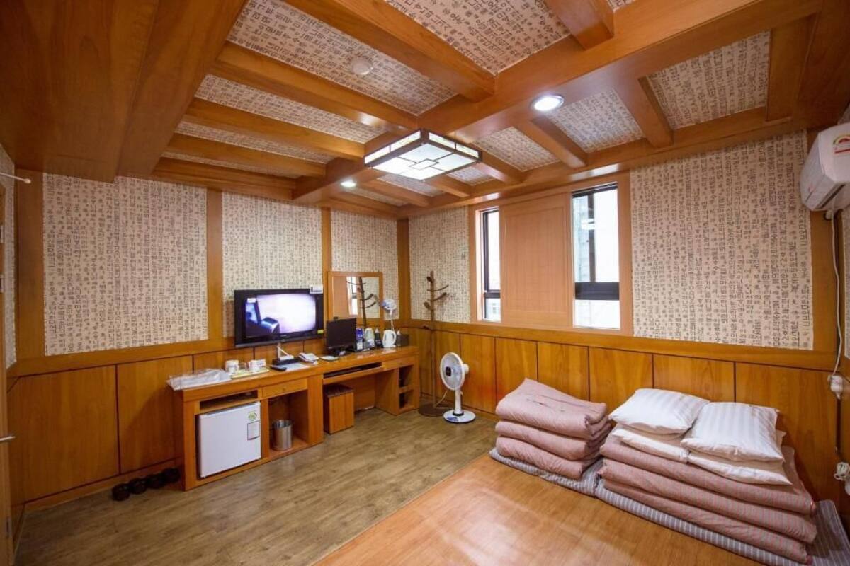A room with wooden walls and ceiling, a television, a desk with a computer, a small refrigerator, and a bed with pillows.