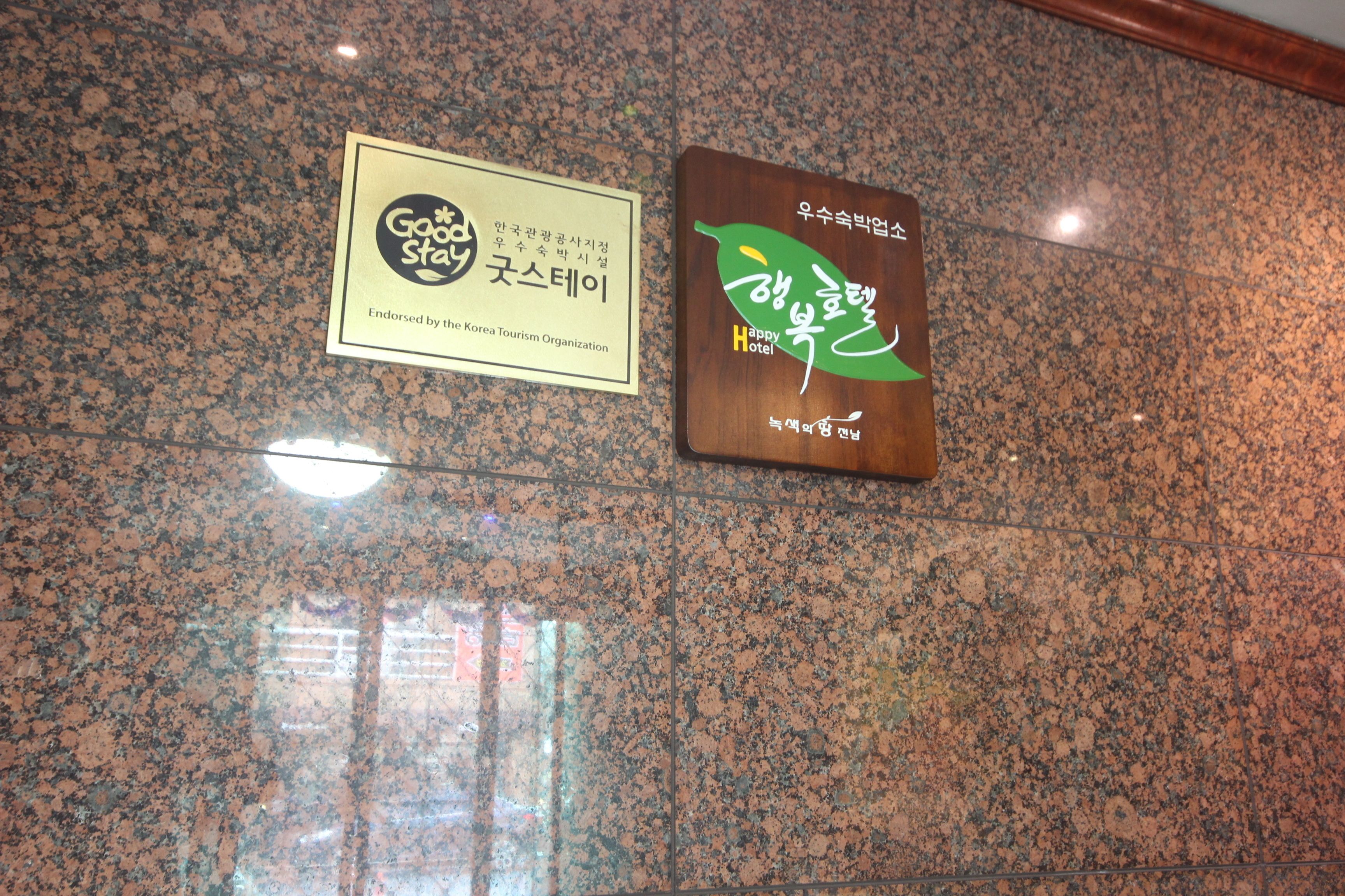 Zwei Schilder an einer Granitmauer: eines ist ein 'Go Stay'-Schild, das vom Korea Tourism Organization empfohlen wird, und das andere ein 'Happy Hotel'-Schild mit koreanischem Text.