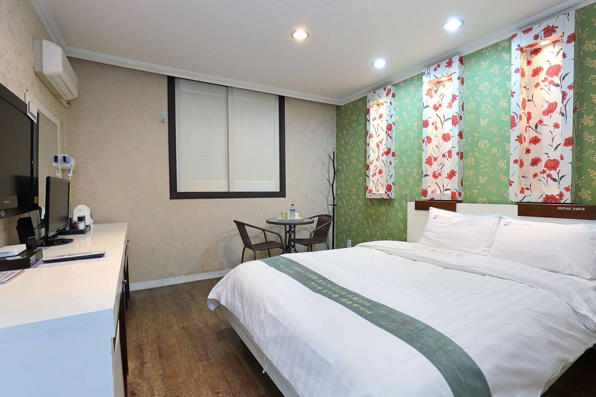 A hotel room with a bed, a desk with a computer, a chair, and a window with blinds.