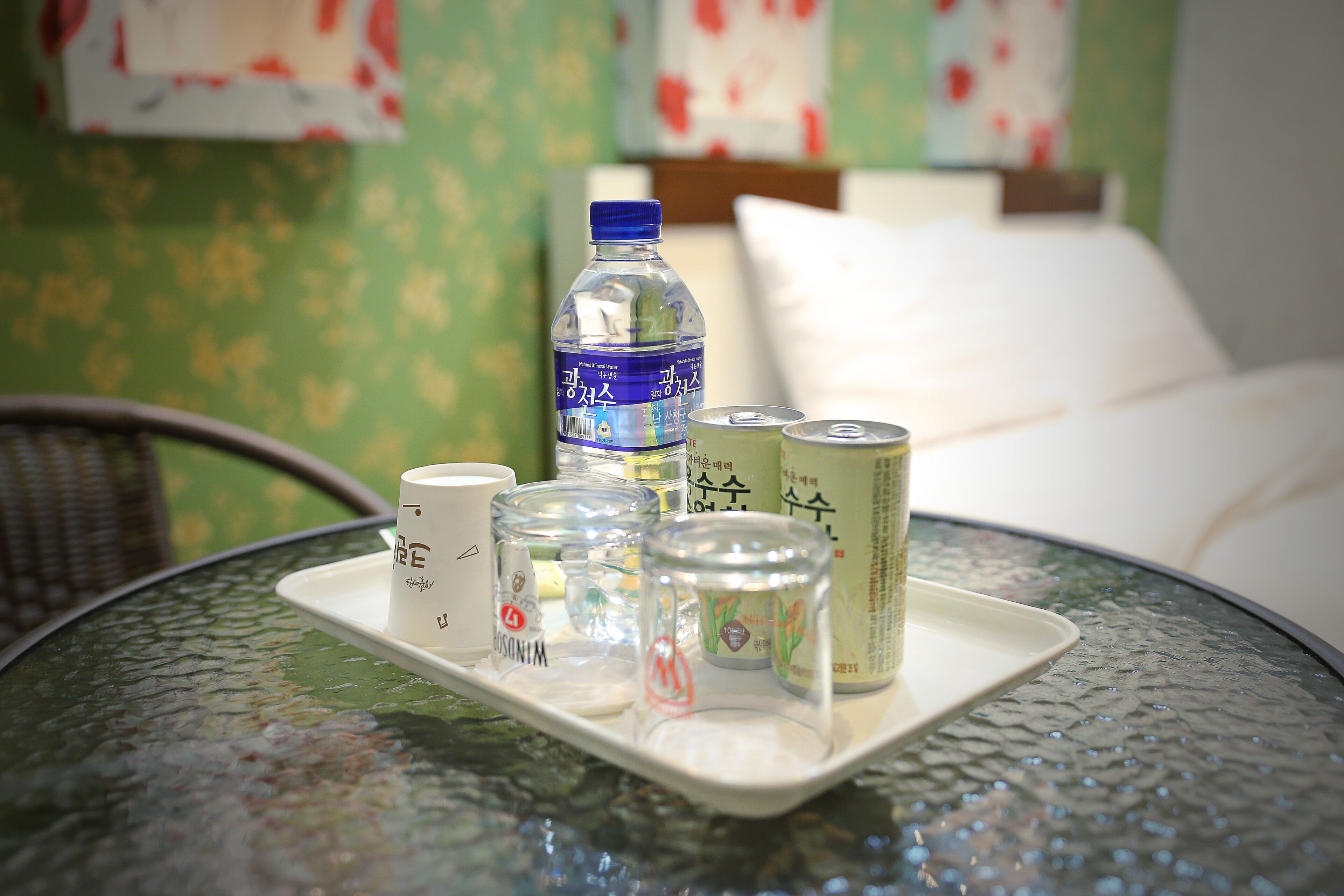 Una bandeja con una botella, latas y una taza sobre una mesa de vidrio en una habitación con papel tapiz floral.