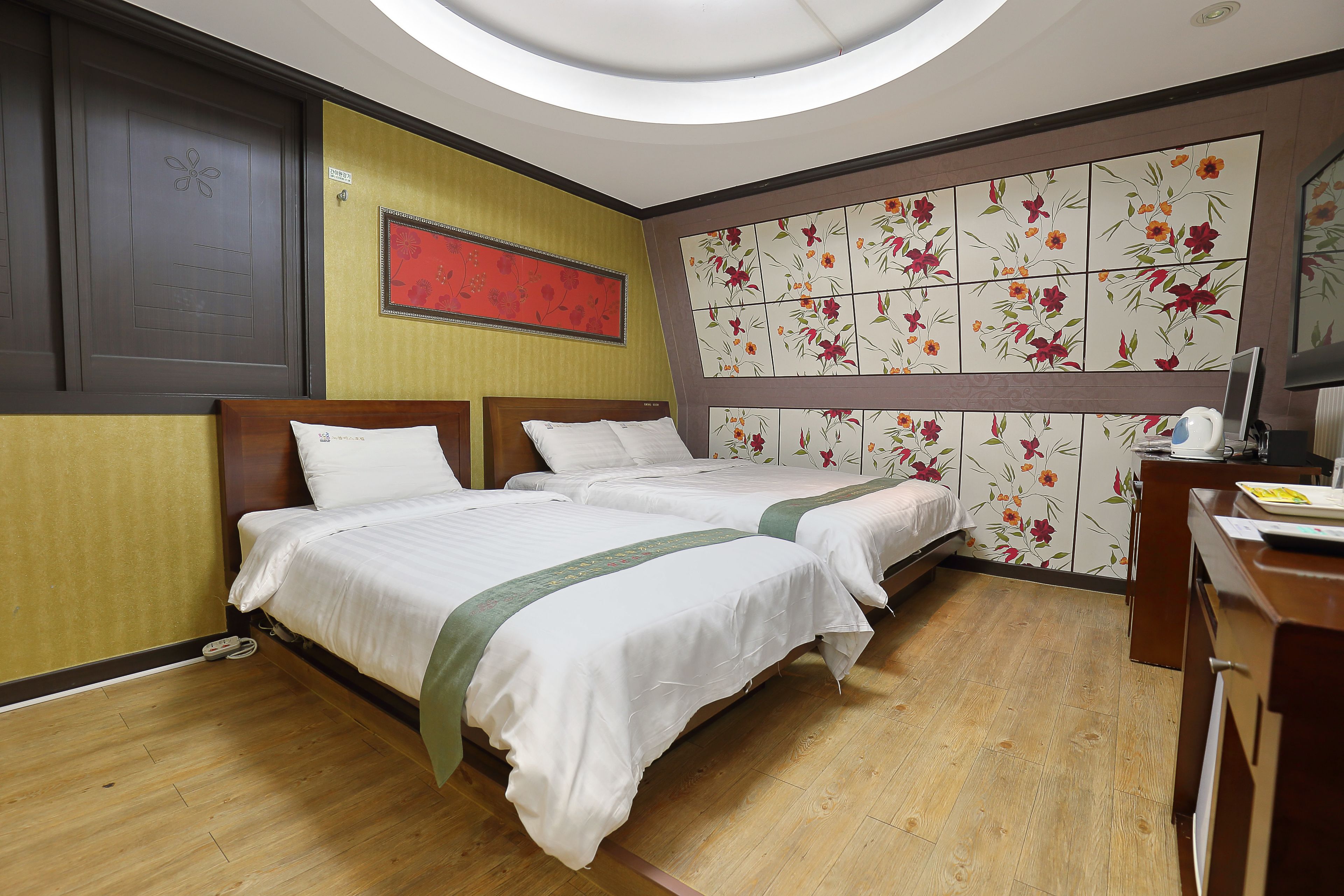 A hotel room with two beds, wooden flooring, a desk with a chair, and a decorative wall with floral tiles.