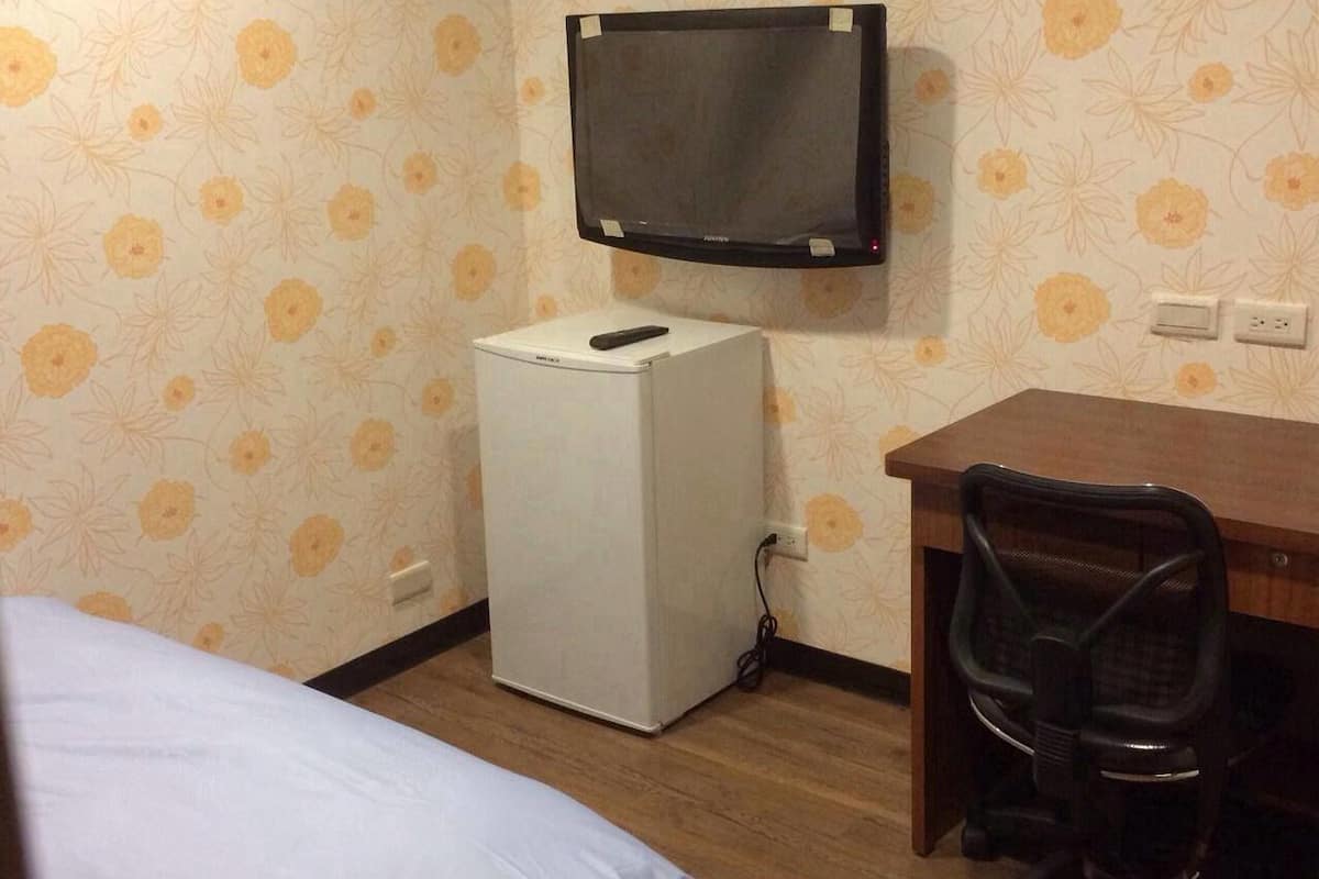Quarto pequeno com uma cama, escrivaninha, cadeira, televisão na parede e geladeira.