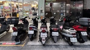 En række parkerede scootere med synlige nummerplader foran en bygning med et tagtækket tag og en skilt.