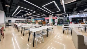 Een moderne cafetaria met witte tafels en stoelen, een schone vloer en een plafond met geometrische verlichtingsarmaturen.