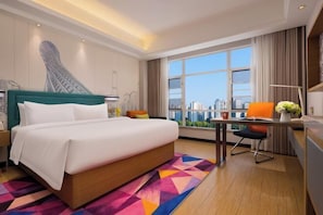 A hotel room with a large bed, a desk with a chair, a view of the city, and a colorful geometric rug.