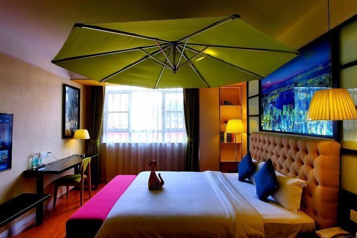 Una habitación de hotel con cama, escritorio, silla, lámpara, un cuadro y una ventana.