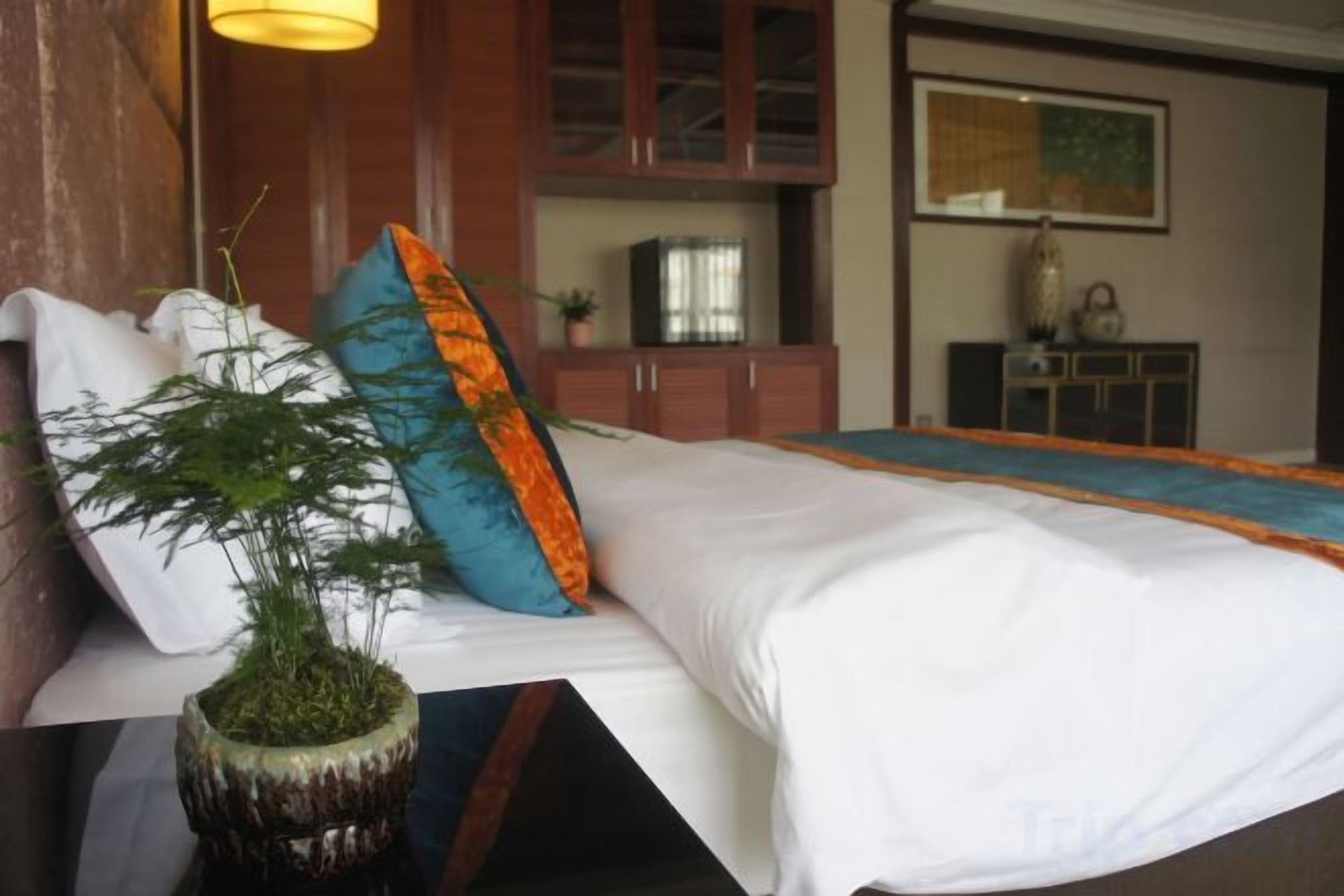 A neatly made bed with a white comforter and a blue and orange pillow, a potted plant, and a wooden cabinet in the background.