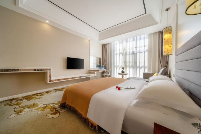 A hotel room with a large bed, a flat-screen TV mounted on the wall, a bedside table, and a window with curtains.