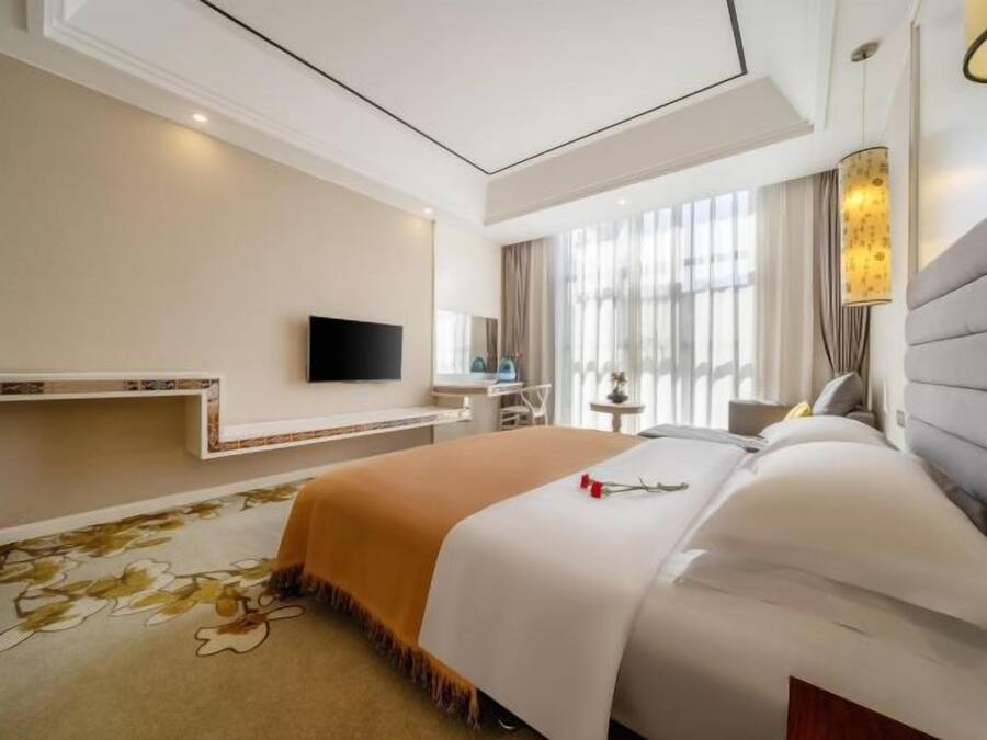 A hotel room with a large bed, a flat-screen TV mounted on the wall, a bedside table, and a window with curtains.