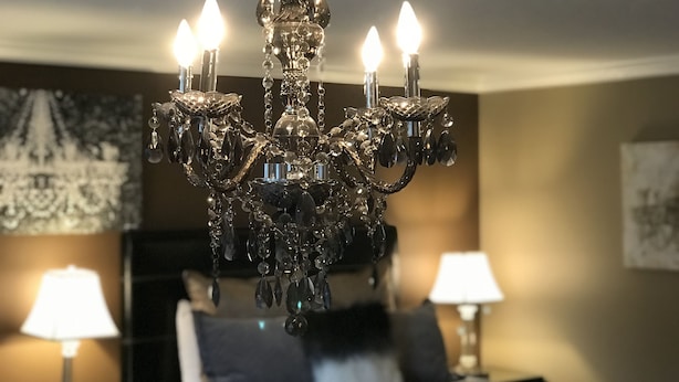 Um candelabro com várias luzes em um quarto com uma cama, duas mesas de cabeceira com abajures, e uma parede com um quadro emoldurado.