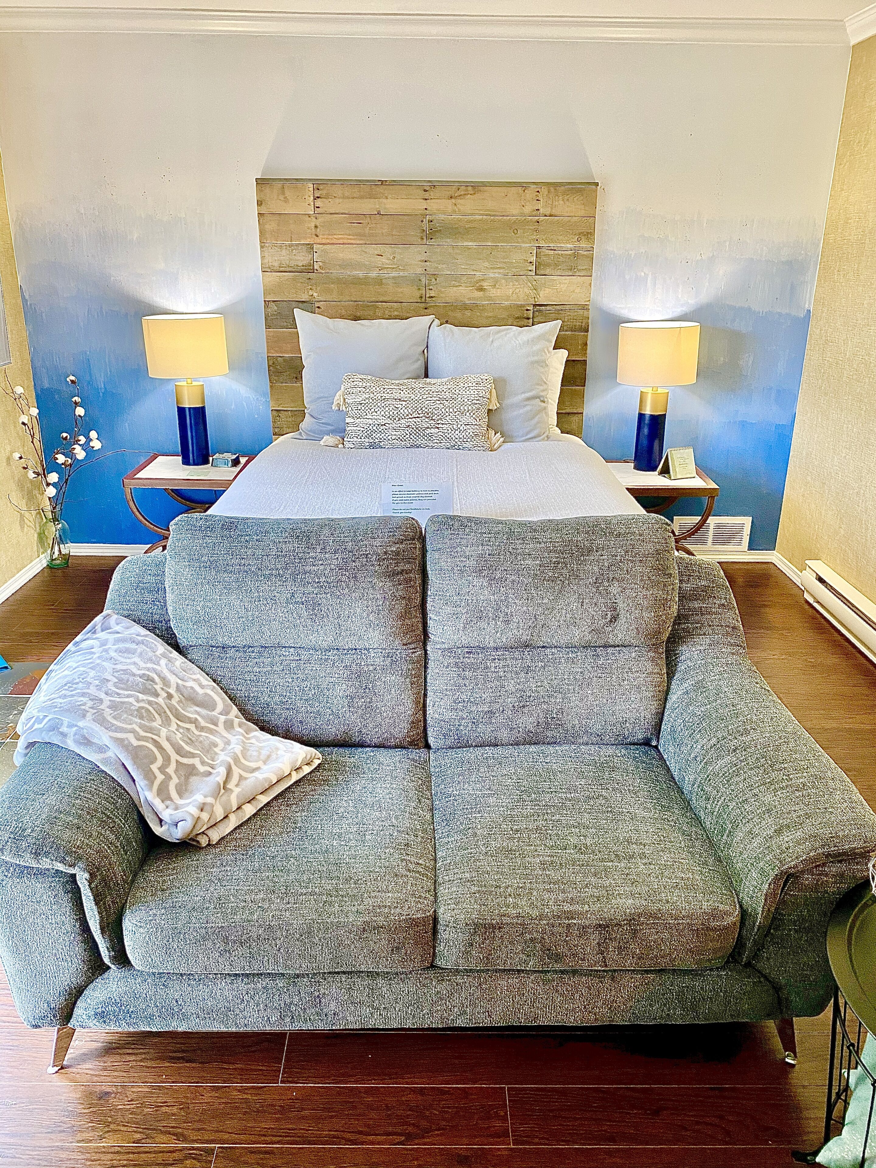 A bedroom with a large bed, two bedside tables with lamps, a grey sofa, and a decorative wall.