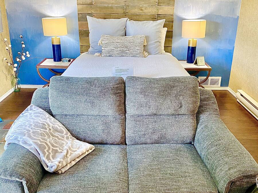 A bedroom with a large bed, two bedside tables with lamps, a grey sofa, and a decorative wall.