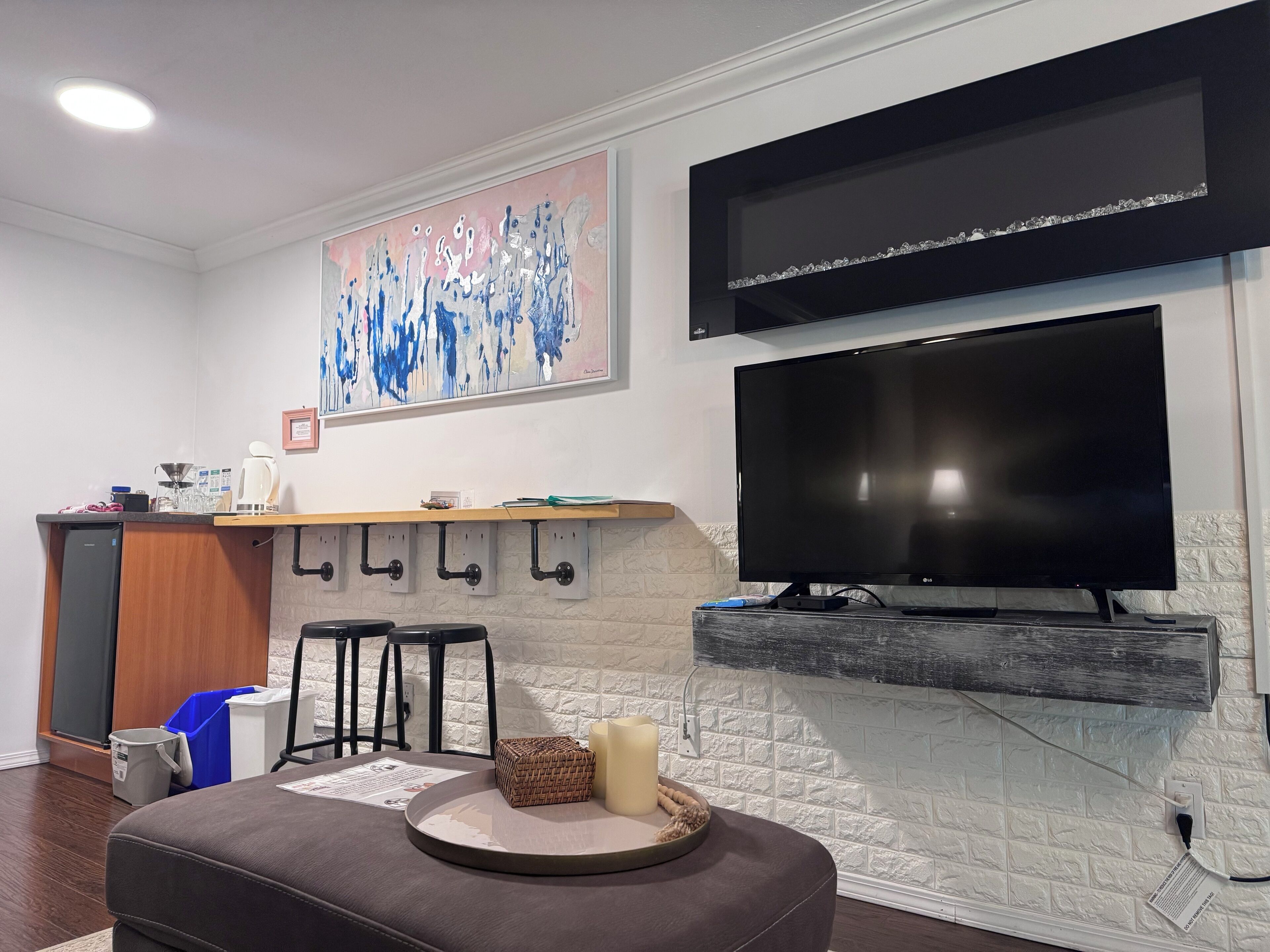 A modern living room with a flat-screen TV mounted on a white brick wall, a kitchen area with a wooden countertop, and a seating area with a cushioned bench.