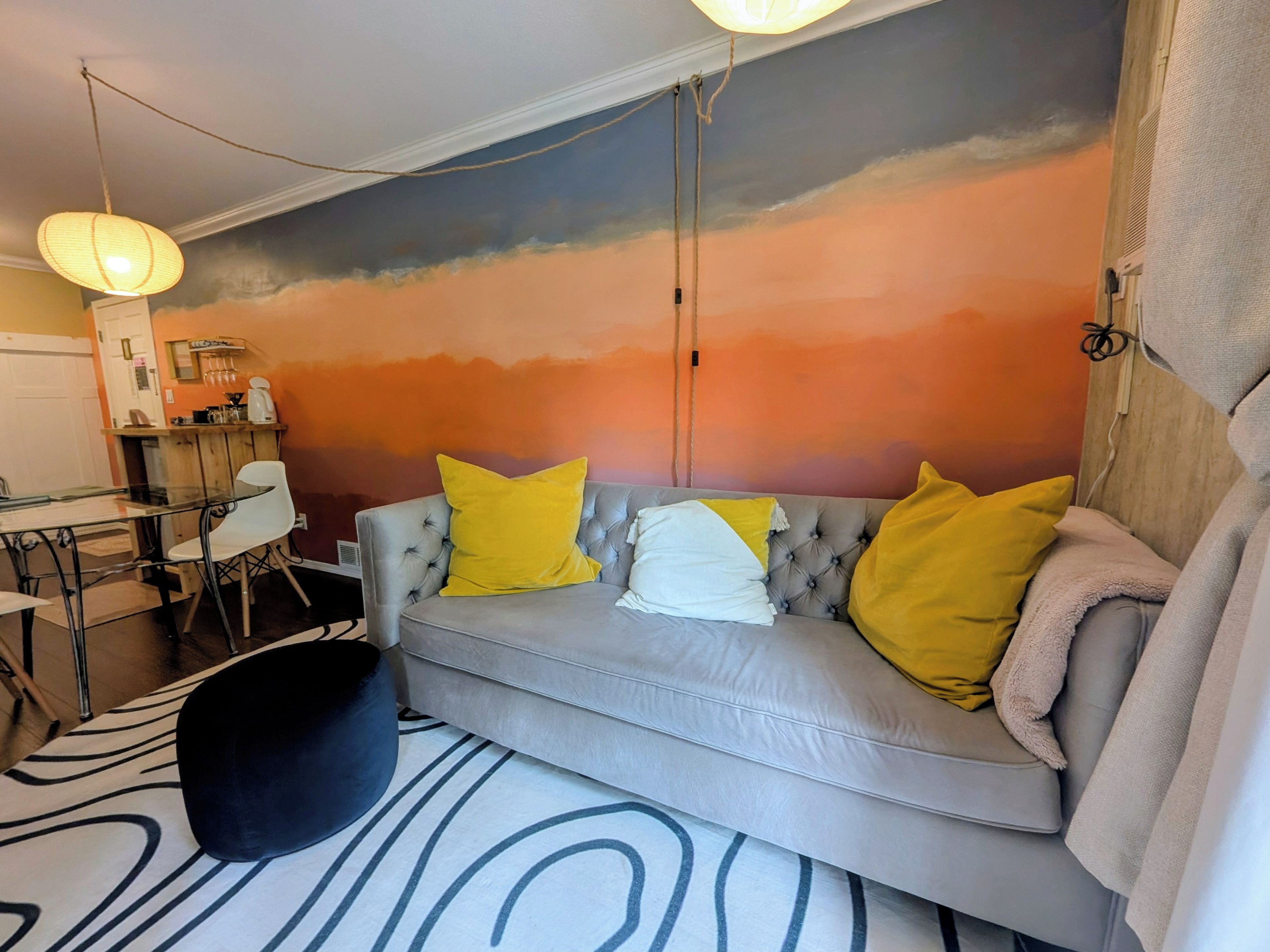 Sala de estar com sofá cinzento, dois almofadas amarelas, um banquinho preto e um tapete estampado.