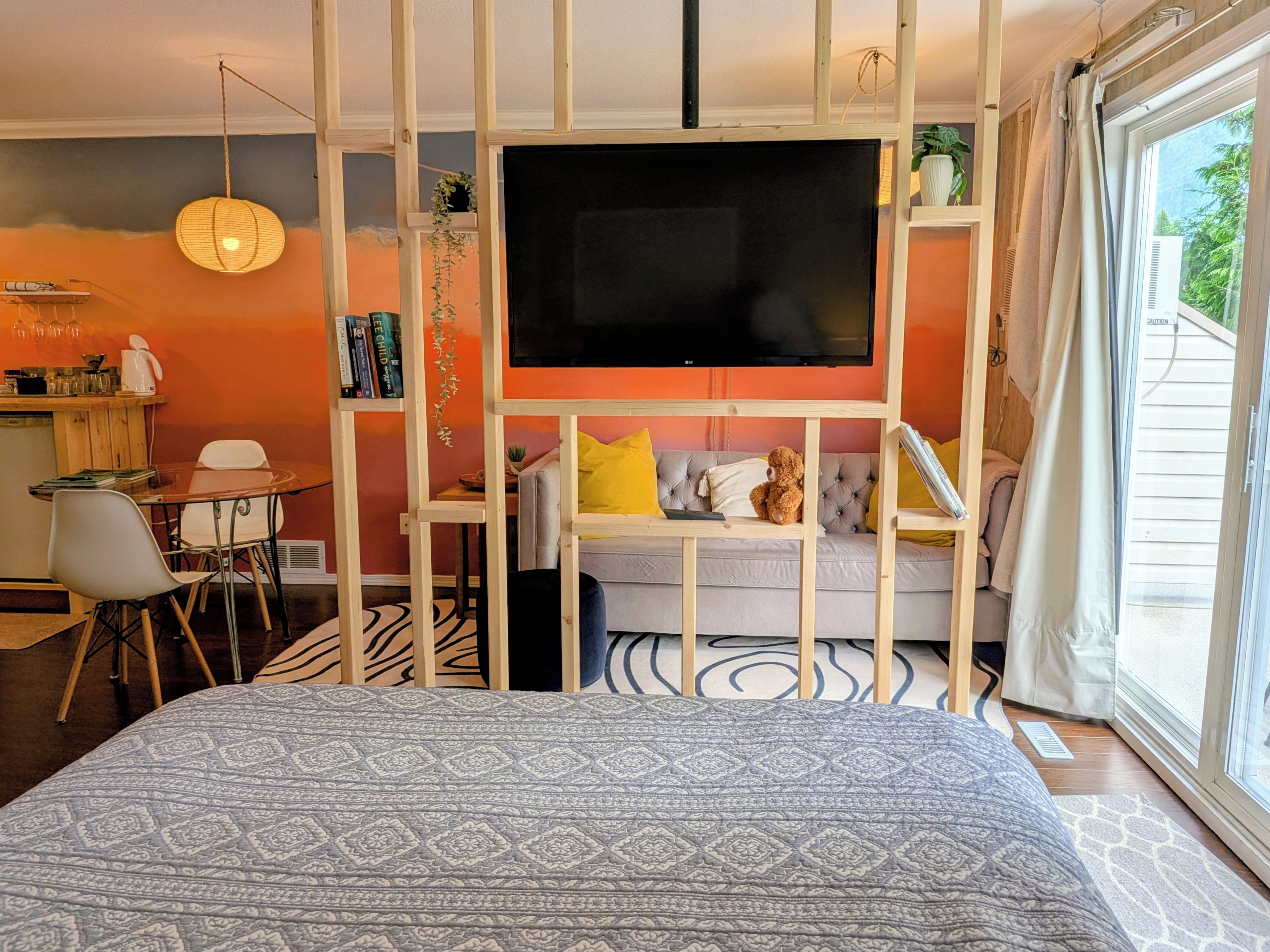 A modern bedroom with a large bed, a TV mounted on a wooden partition, a dining area with a table and chairs, and a view of the outdoors through a window.