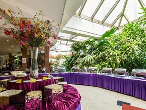 A banquet hall with a high ceiling and skylights, featuring a central floral arrangement, a buffet setup with food service stations, and lush green plants. - The Twenty First Century Hotel - Beijing (Beijing)