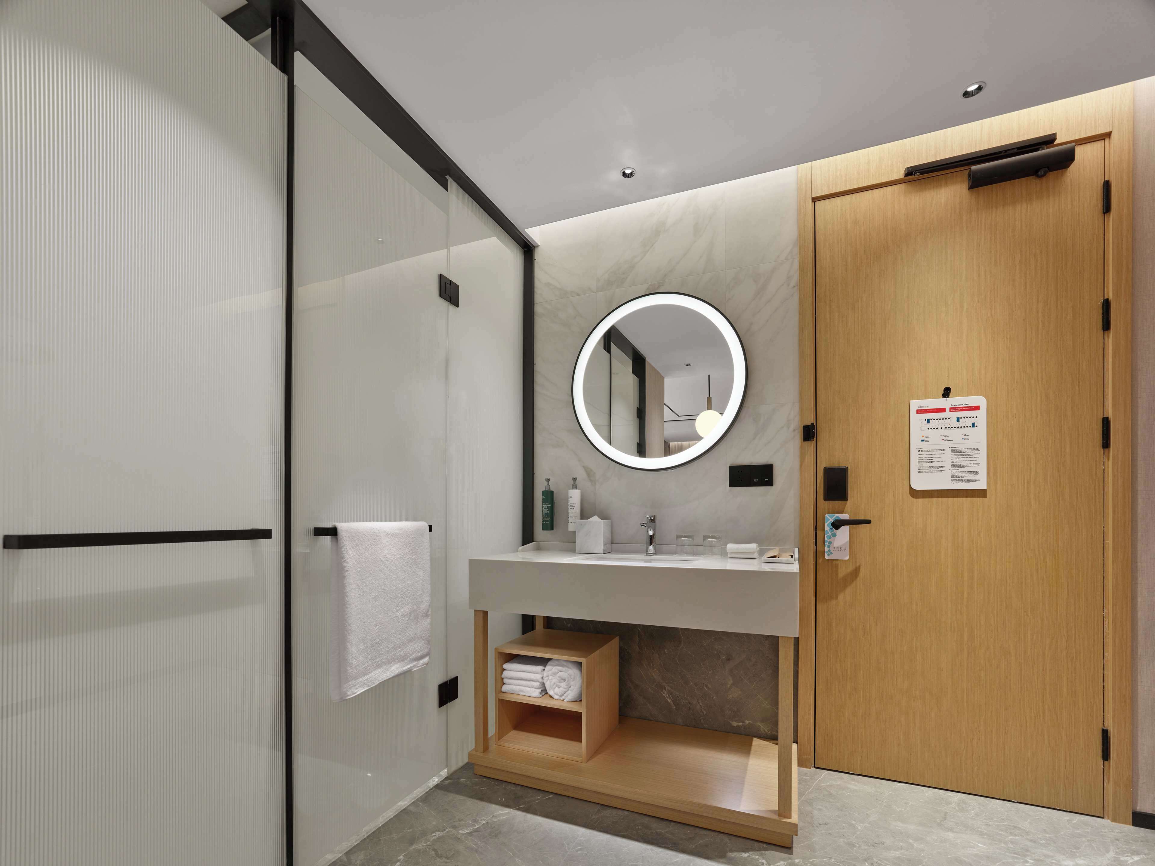A modern bathroom with a glass shower enclosure, a wooden vanity with a marble countertop, a round mirror, and a wooden door with a sign.