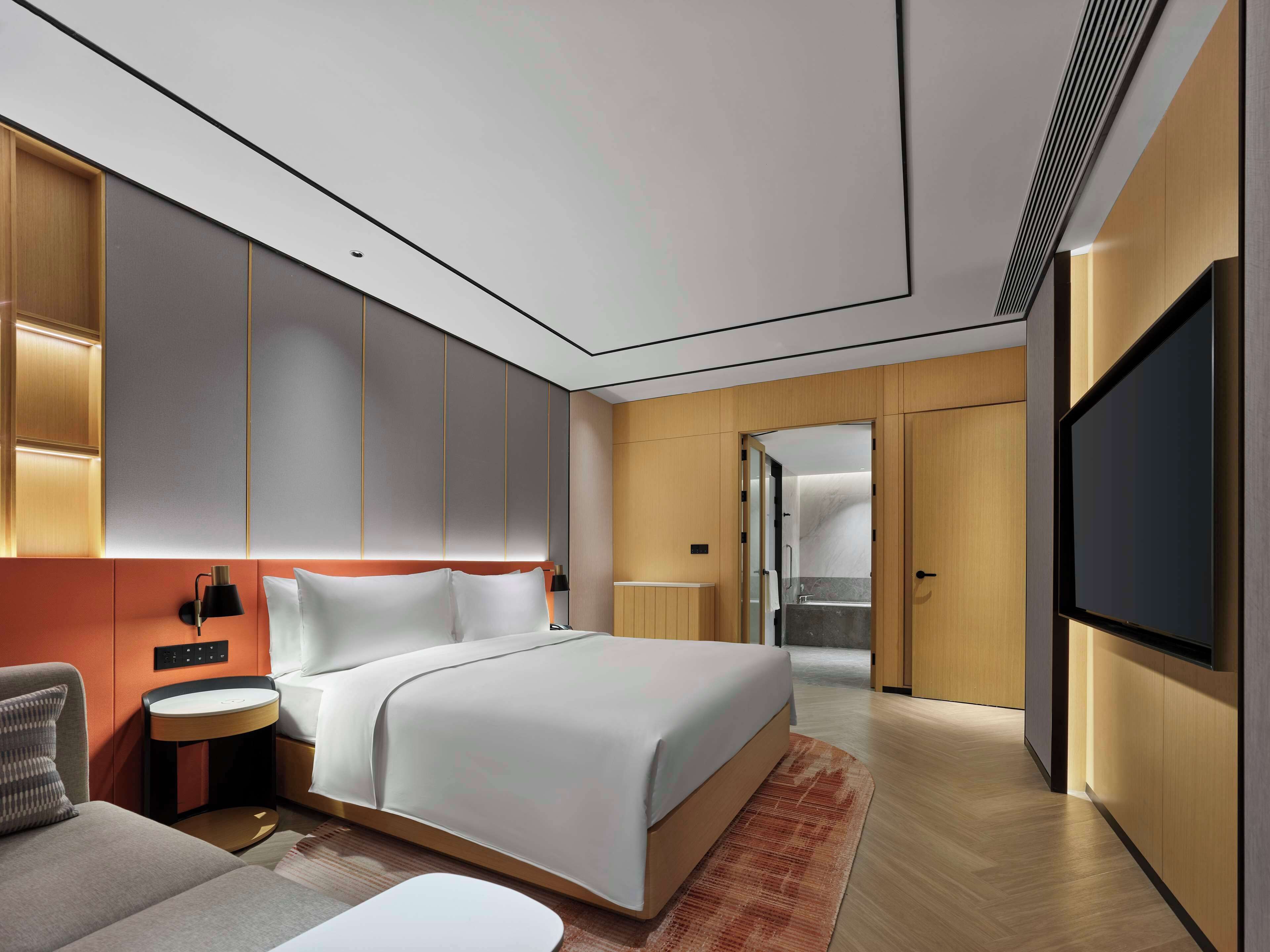 A modern hotel room with a large bed, a bedside table, a TV, and a bathroom visible through an open door.