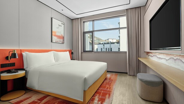 A modern hotel room with a bed, a desk, a TV, and a window with a view of buildings. - Hilton Garden Inn Huangshan Scenic Area South Gate (Huangshan)