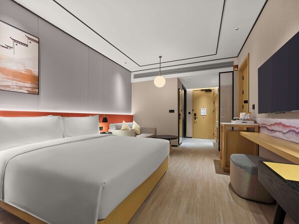 A modern hotel room with a large bed, a desk with a flat-screen TV, and a seating area with a patterned ottoman. - Hilton Garden Inn Huangshan Scenic Area South Gate (Huangshan)