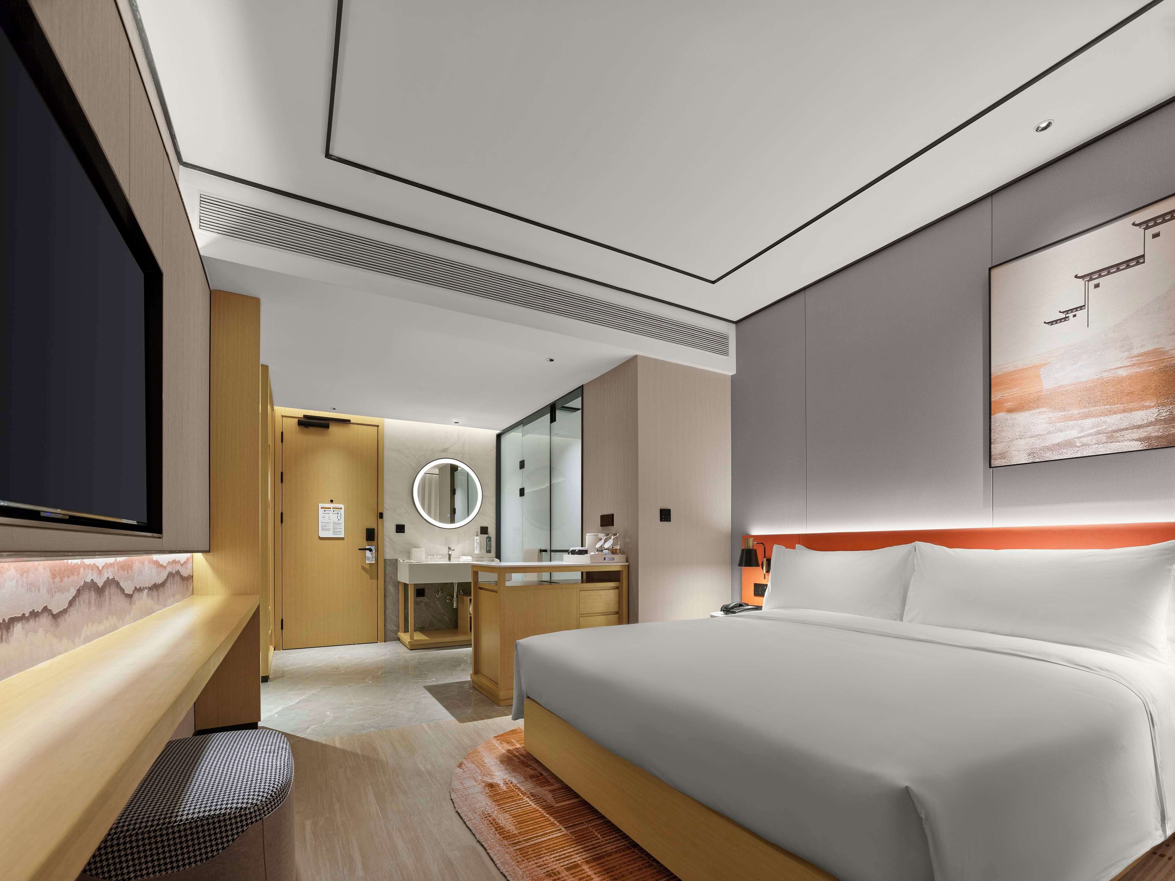A modern hotel room with a large bed, a desk, a mirror, and a television.