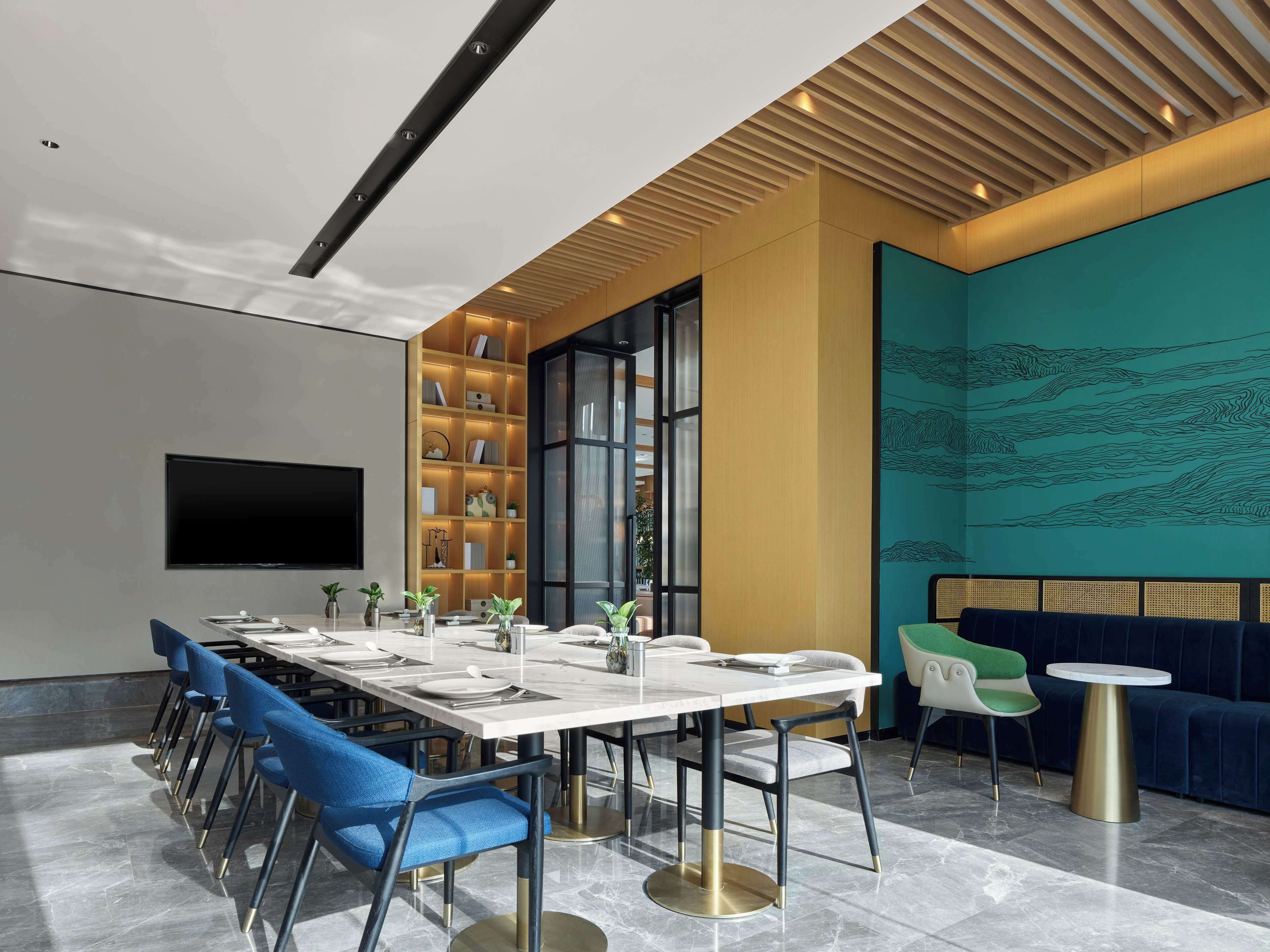 A modern dining area with a long table, blue chairs, a sofa, and a small round table.