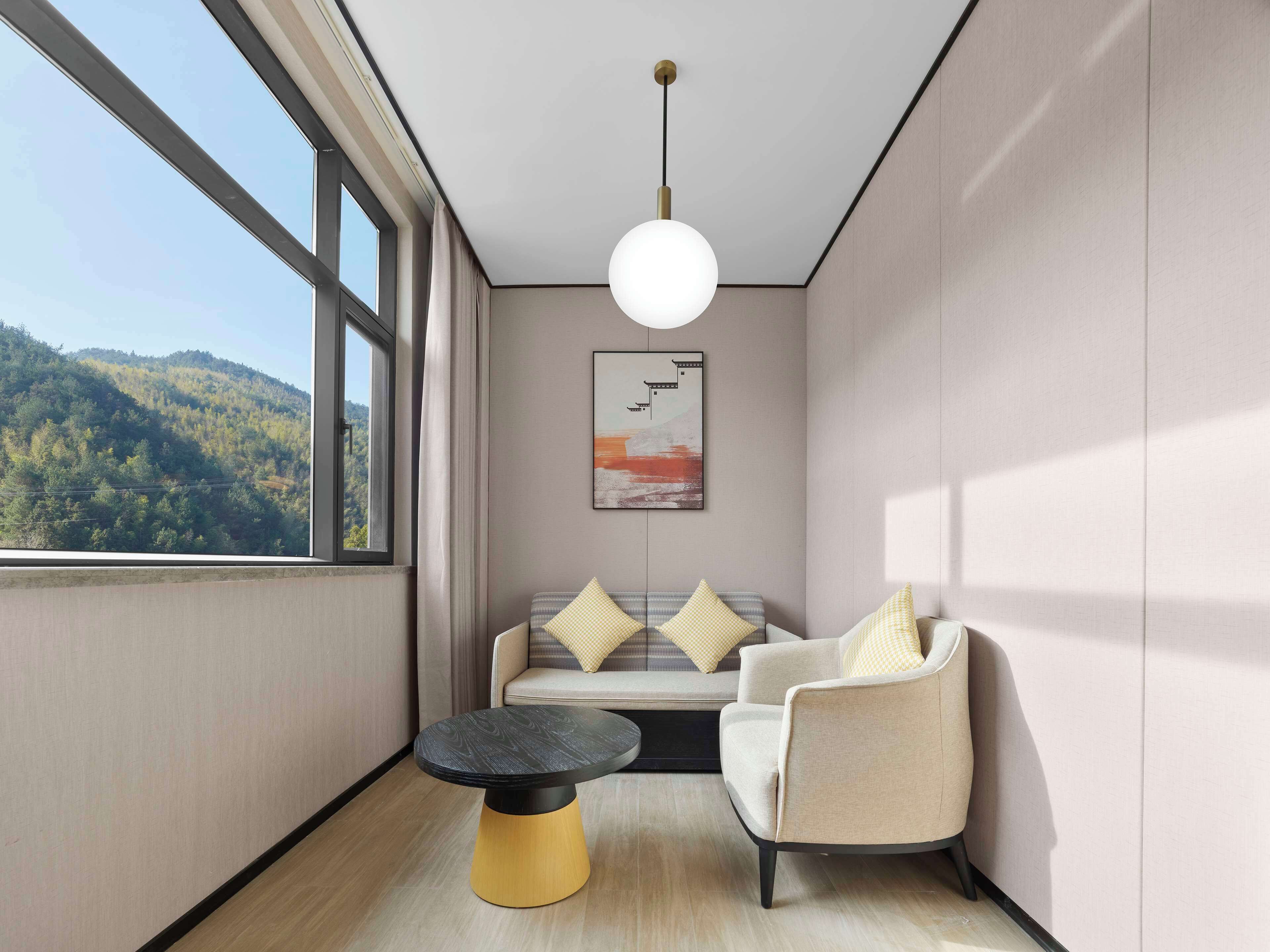 A modern room with a large window, a round table, a sofa with cushions, and a hanging light fixture.