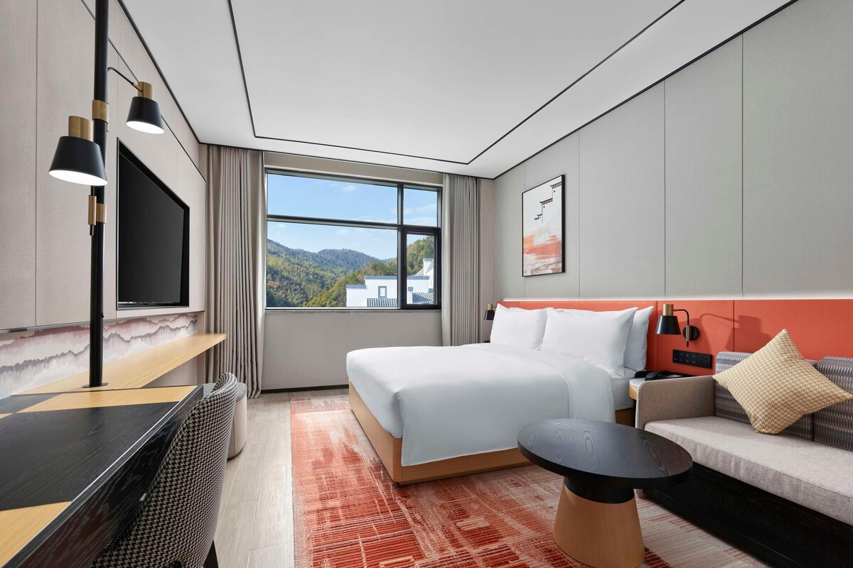 Habitación de hotel con cama, sofá, televisión, escritorio y ventana con vistas a la montaña.
